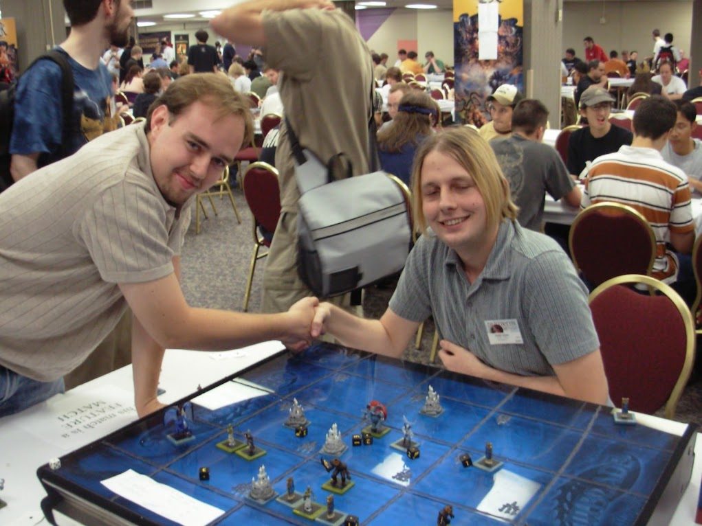 Post-match handshake across a miniatures battle map in a crowded tournament hall